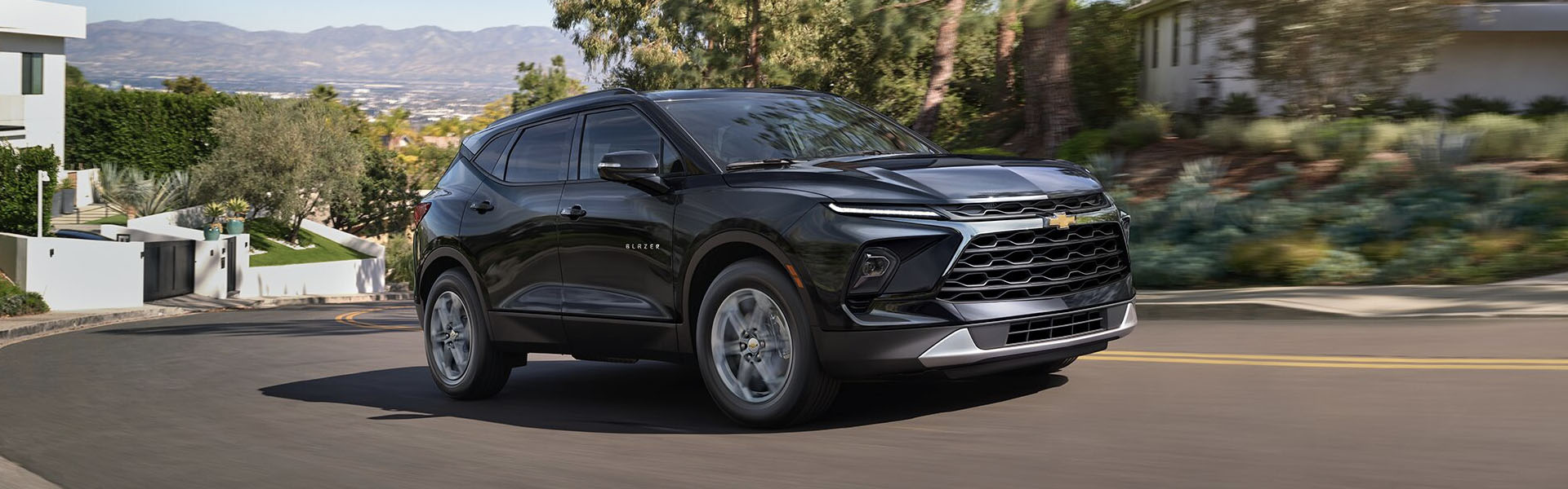 2025 Chevrolet Blazer in black driving on a curved suburban road, shown from a front three-quarter angle with LED headlights, bold grille, and modern SUV styling, surrounded by trees and hillside homes.