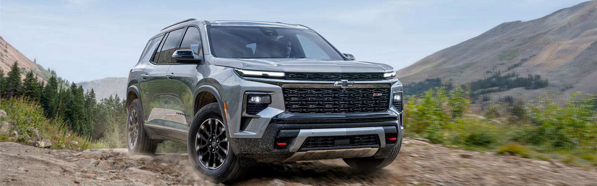 2026 Chevy Traverse silver midsize SUV driving on a rugged mountain trail with scenic hills in the background