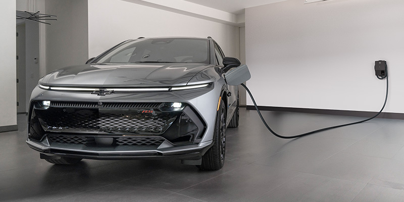 A sleek, silver electric Chevrolet Equinox EV is parked in a modern garage, plugged into a wall-mounted charger. The setting conveys innovation and eco-friendliness.