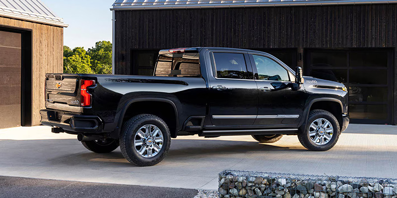 A black pickup Chevrolet Silverado HD truck is parked on a light-colored driveway near a modern wooden building. The truck's polished surface reflects sunlight, conveying a sleek, robust appearance.