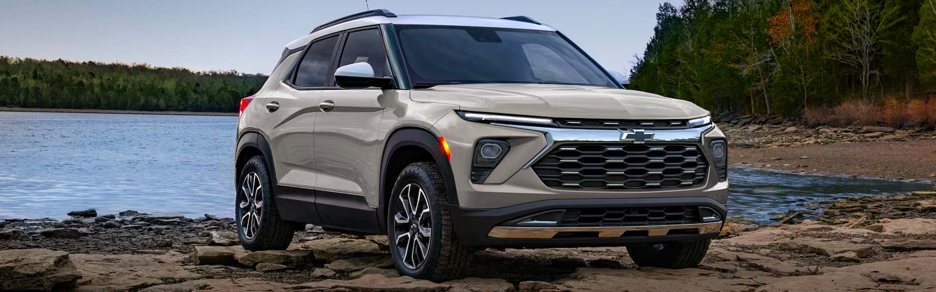 A beige 2026 Chevrolet Trailblazer parked on rocky terrain by a lake, surrounded by greenery.